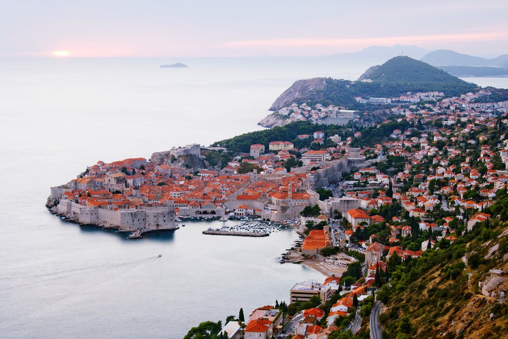 The historic old city of Dubrovnik with its walls, red tiled roofs, harbor, and sea at sunset.