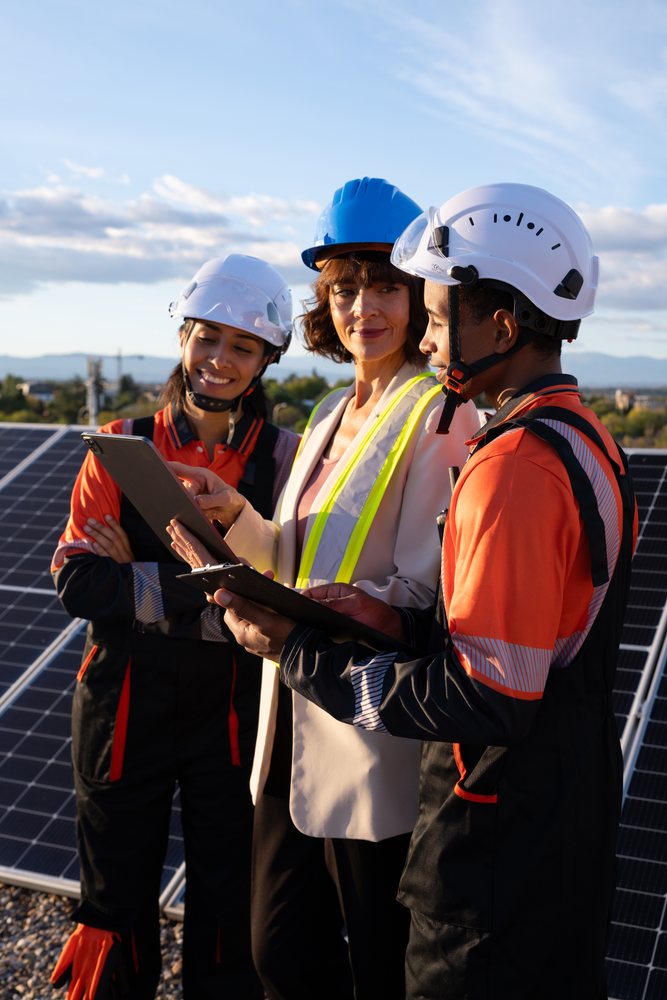 Three engineers wearing safety helmets and vests inspect solar panels outdoors, discussing solar energy under daylight, Madrid, Spain.