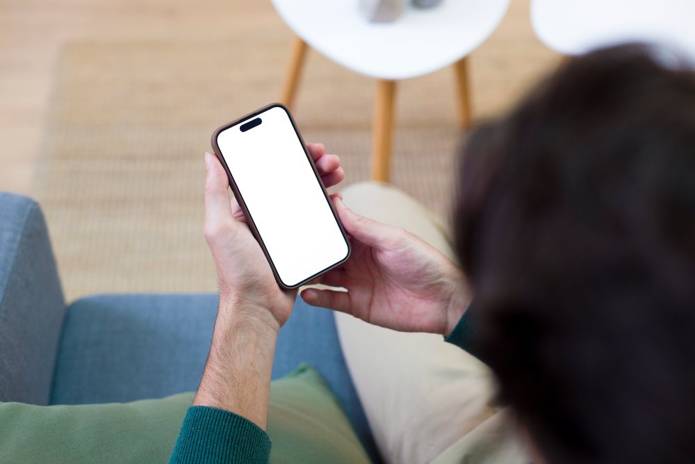 Man sits on a couch in a home setting holding a smartphone with a blank screen, Technology, young adult.
