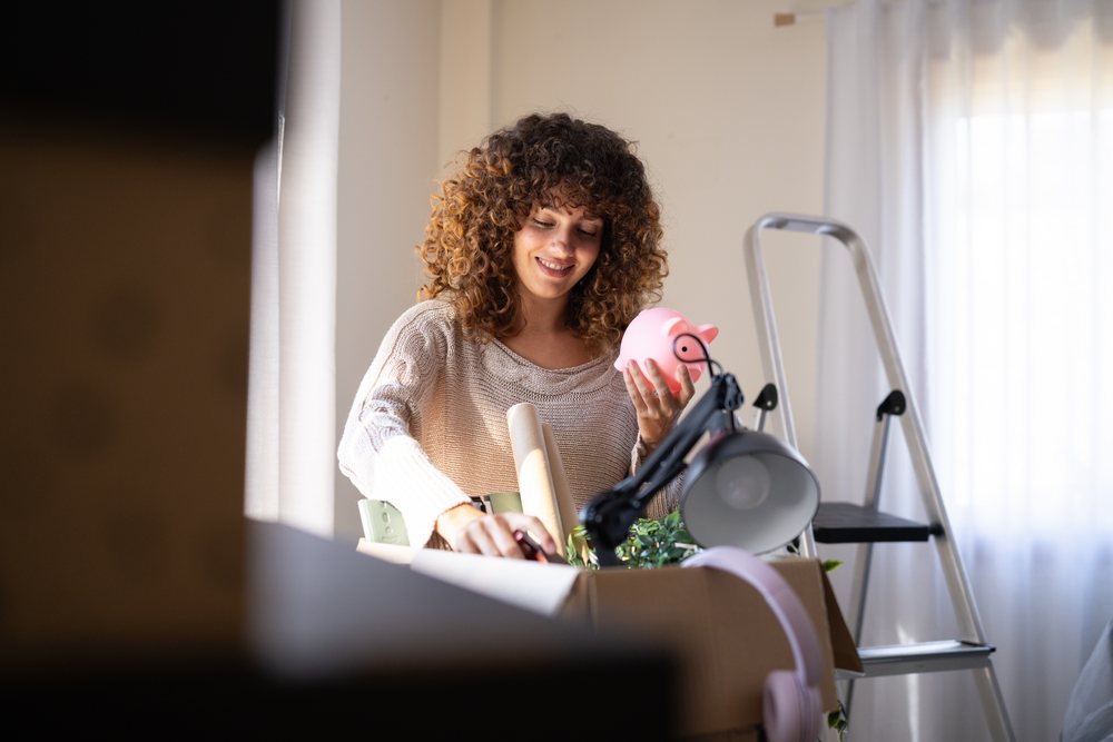 Woman smiling as she unpacks a cardboard box containing a pink piggy bank, desk lamp, plant, and headphones while moving indoors, Spain.