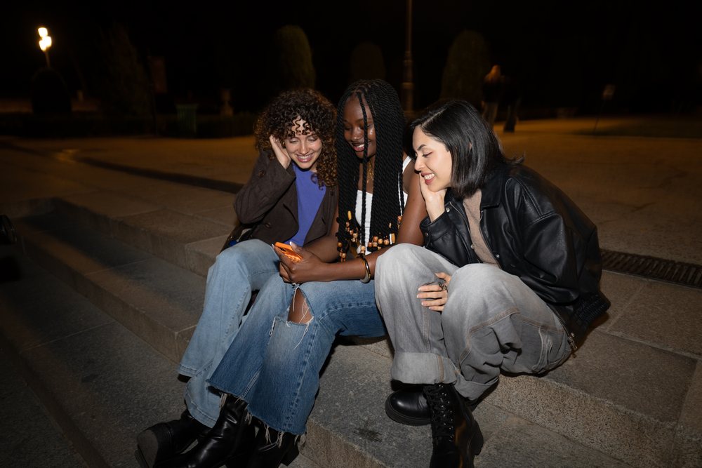 Three women sitting on stone steps outdoors at night in an urban setting, smiling and watching a smartphone together, Madrid, Spain.