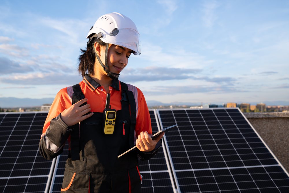 Woman solar panel technician inspects solar panels outdoors using a tablet, wearing safety gear under blue sky in an urban environment, Madrid, Spain.