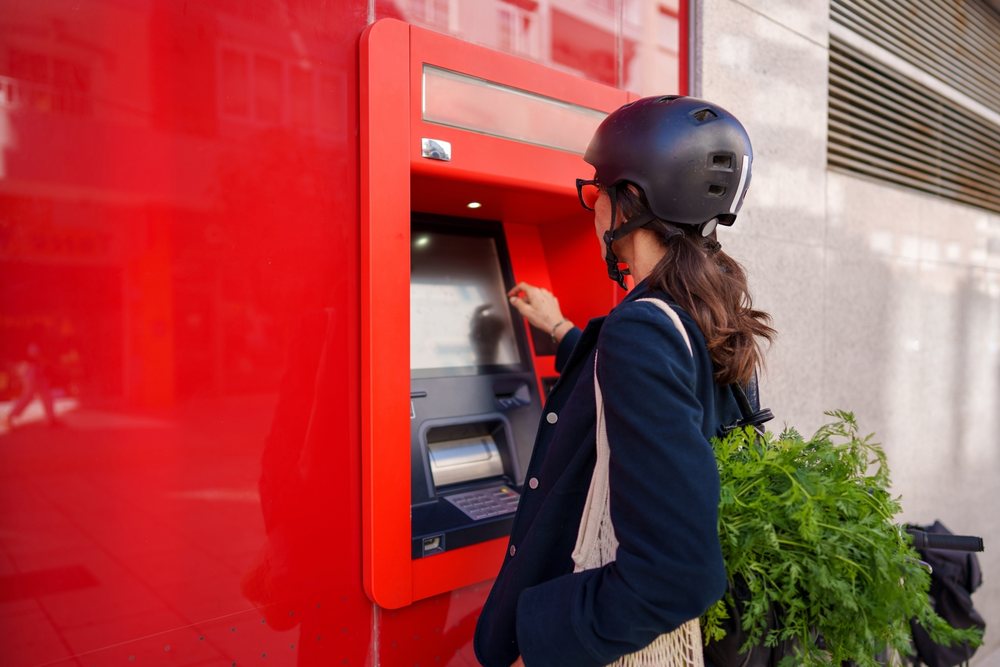 Woman wearing helmet uses outdoor ATM with bicycle and mesh shopping bag of fresh greens in urban Málaga setting.