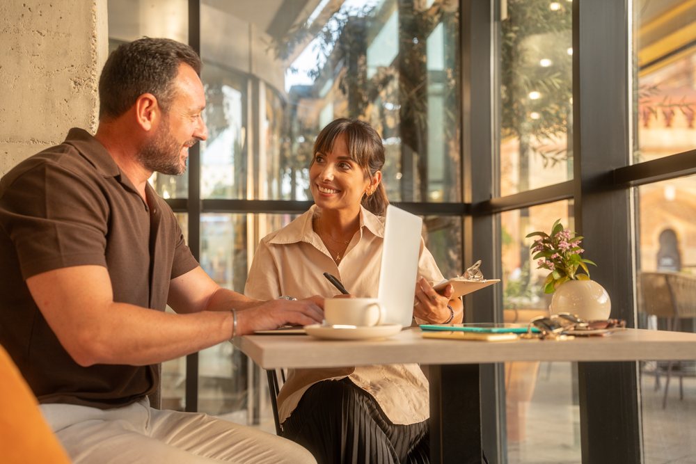 Two coworkers collaborate on a business project in a bright office with laptop, tablet, and coffee cup, Seville, Andalucia.