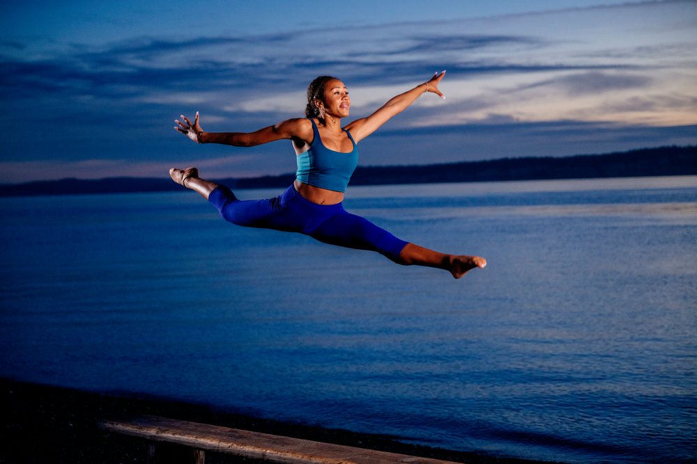 Woman performing a yoga acrobatic leap with arms extended outdoors by the ocean at sunset, USA.