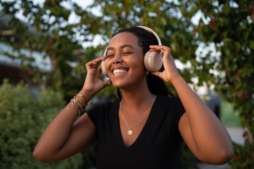 A smiling woman with headphones enjoys music outdoors with greenery in the background, urban, Santiago De Compostela, Spain.