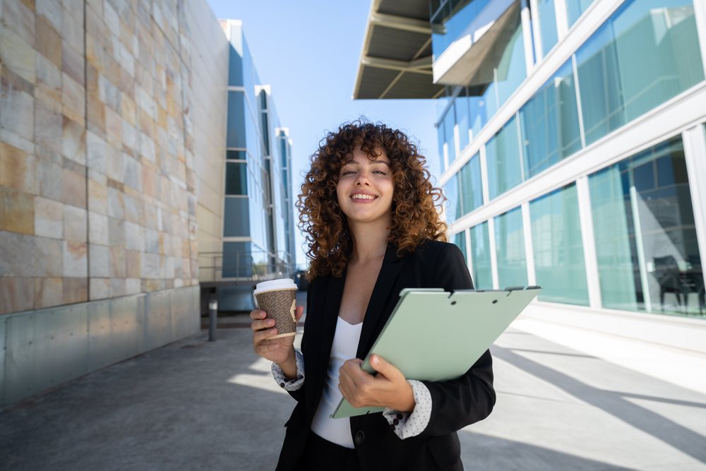 Businesswoman holding a coffee cup and clipboard outdoors near modern office buildings, traveling executive, Spain.