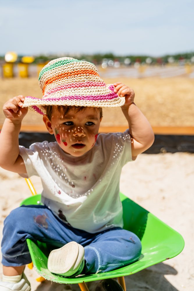 Toddler with red face paint sitting inside a green wheelbarrow outdoors wearing a colorful woven straw hat under bright sunlight, kids, Spain.