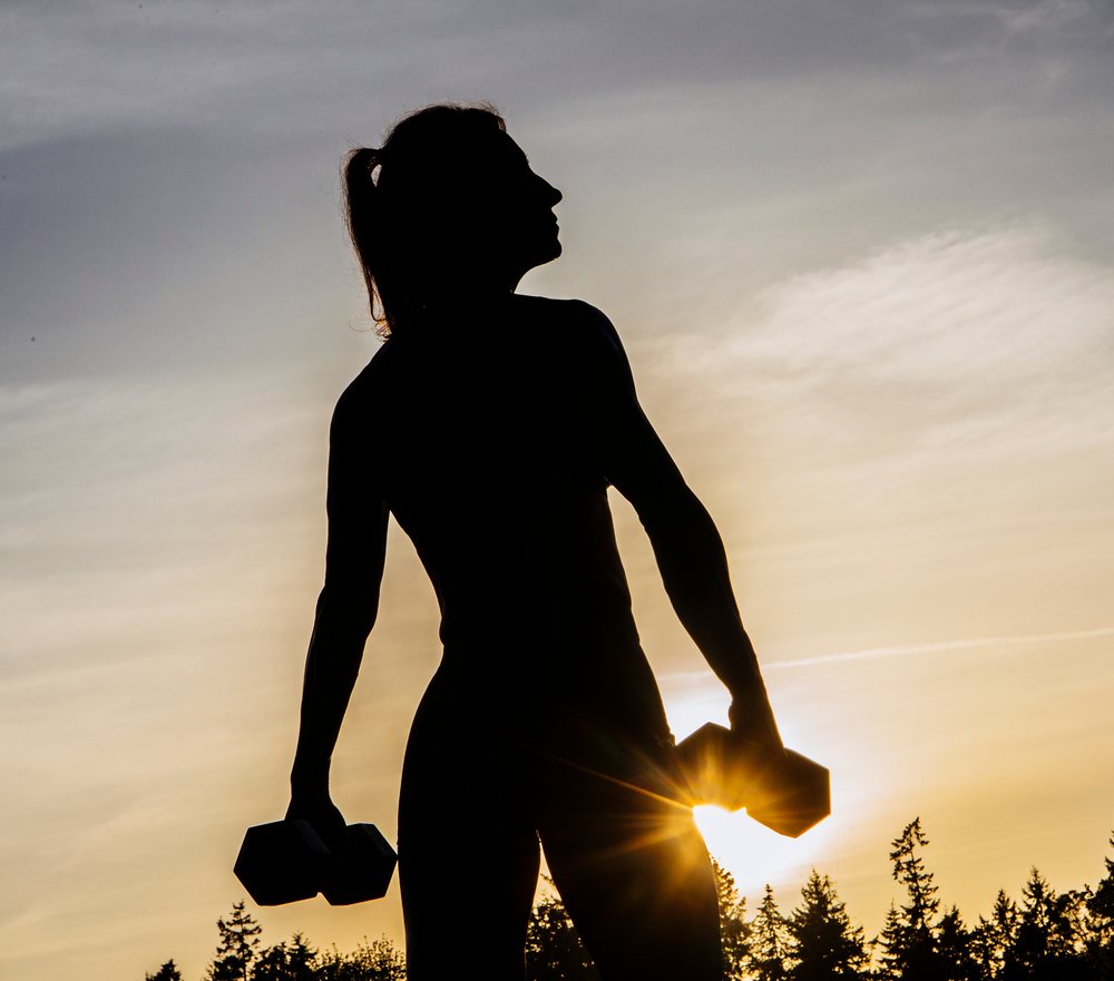 Silhouette of a woman lifting dumbbells outdoors at sunset in USA practicing calisthenics with trees and sky in background.