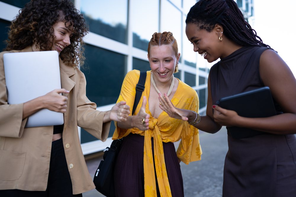 Three friends in casual clothing play a hand game outdoors in an urban area, smiling and holding a laptop and tablet, Spain.