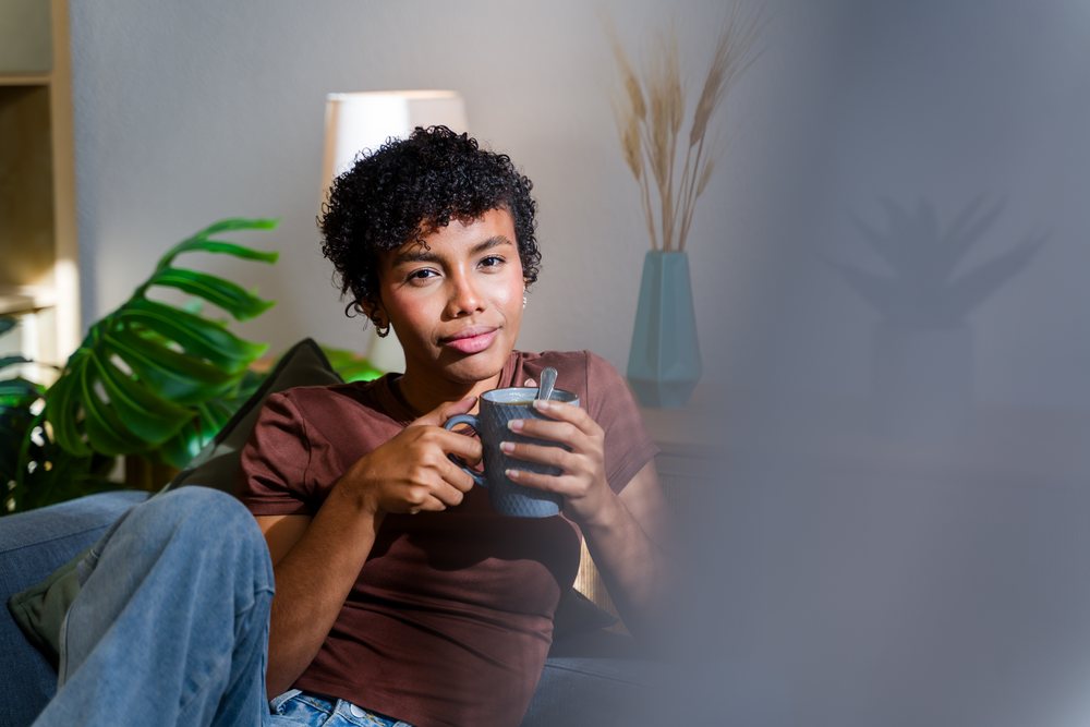 Woman sits indoors on a sofa holding a cup, with plants, a lamp, and a pillow visible behind her, Spain.