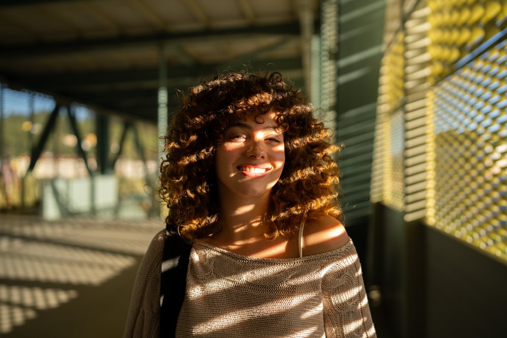 Woman with curly hair smiling in a sunlit outdoor corridor with striped shadows, home story, influencer, Spain.