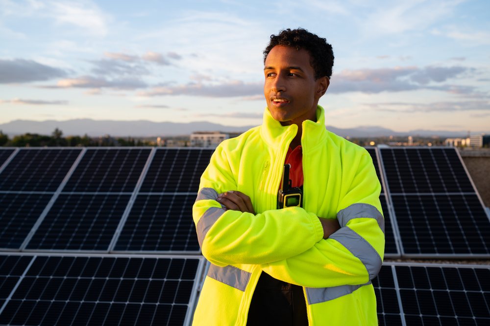 Man wearing bright yellow high-visibility jacket stands with crossed arms by rows of solar panels at sunset outdoors, Madrid, Spain.