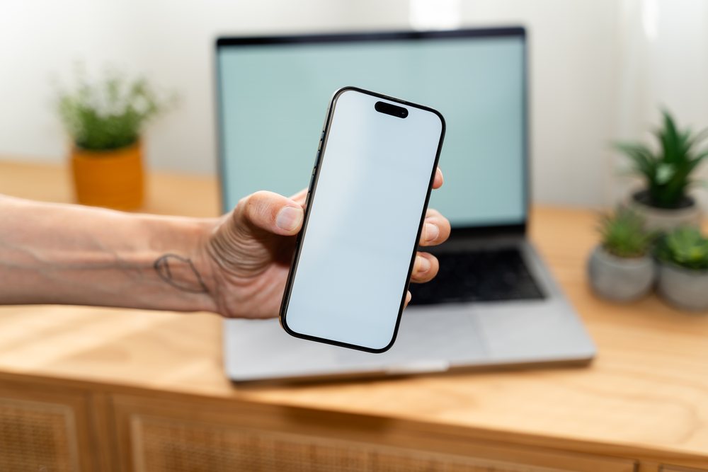 Smartphone with blank screen held by left hand showing tattoo, with laptop and potted plants on wooden office desk, mock up, white screen, Spain.
