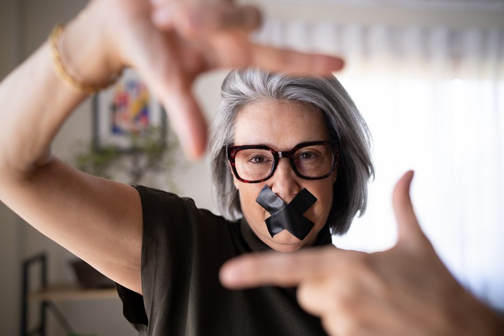 Woman with gray hair and glasses indoors, framing her face with hands and black tape over her mouth, Spain.