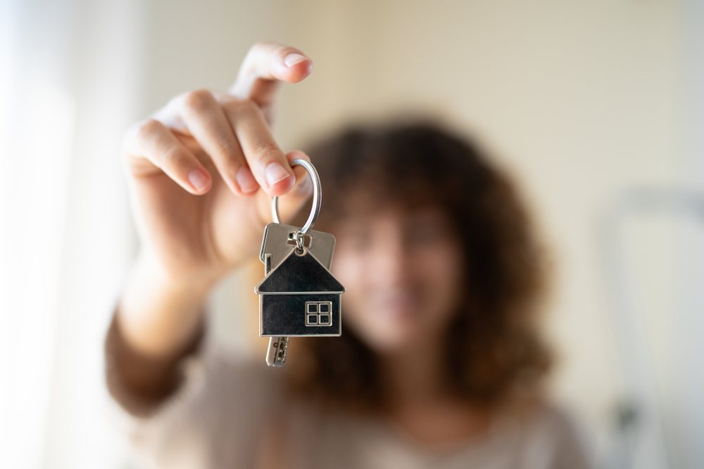 Silver keys with a metal house-shaped keychain held indoors by a person with blurred face in background, moving, Spain.