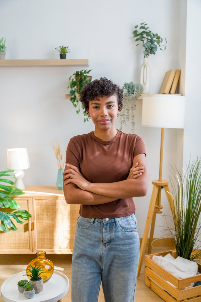 Young woman with crossed arms stands inside a modern living room decorated with plants and wooden furniture.