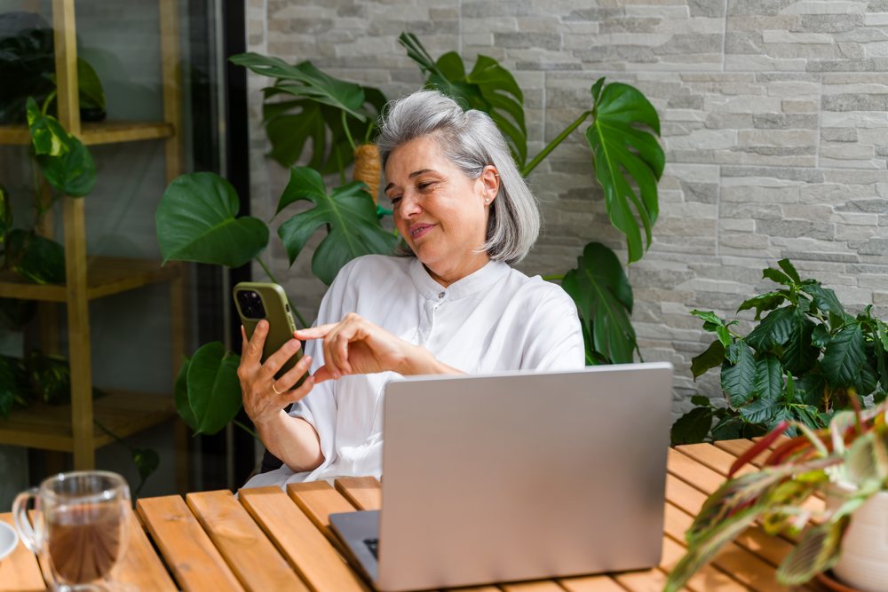 Woman using smartphone while sitting at a wooden table with a laptop and surrounded by indoor plants, Spain.