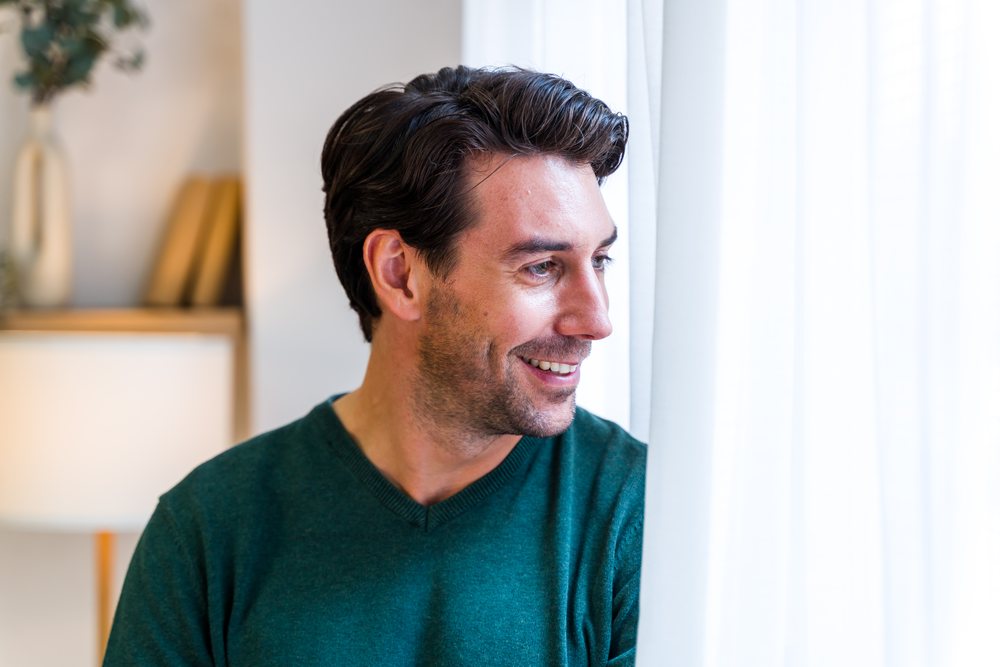 Man with dark hair and stubble smiling inside home looking out window past white sheer curtains near lamp and books, Spain.