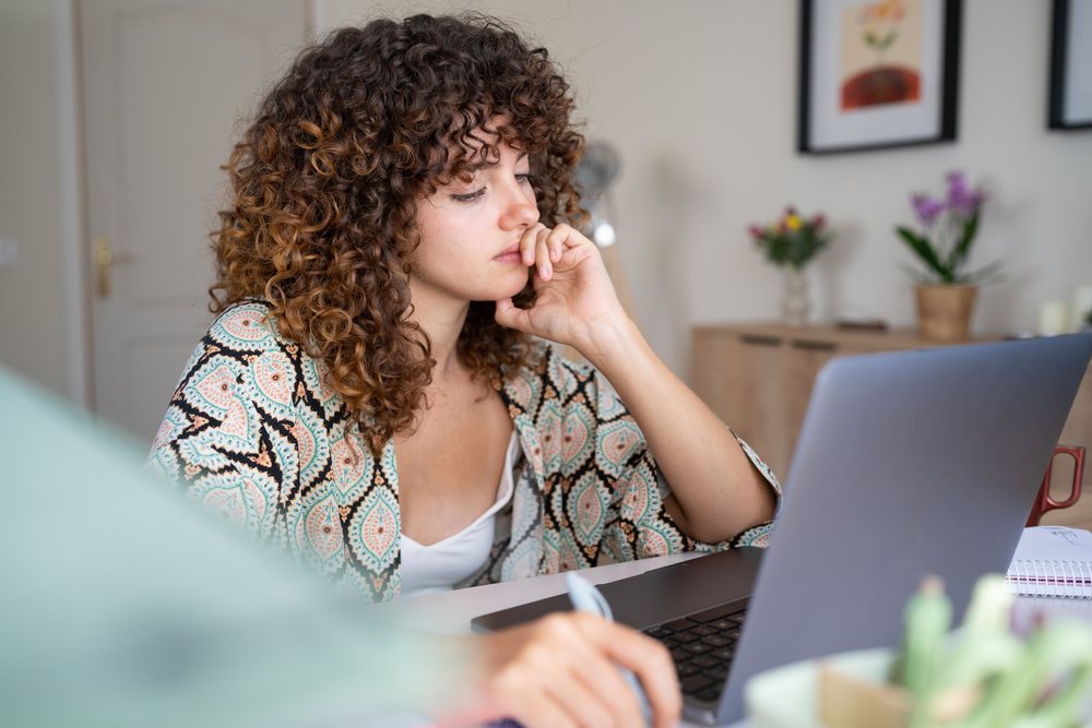 Girl with curly hair concentrates while writing in a notebook and using a laptop at home, Spain.