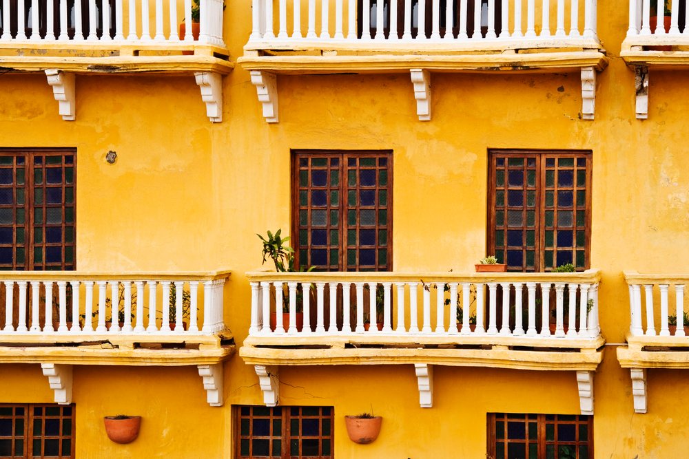 Yellow painted colonial building facade with white balconies, wooden framed colored glass windows, and terracotta pots with plants, Cartagena, Columbia.