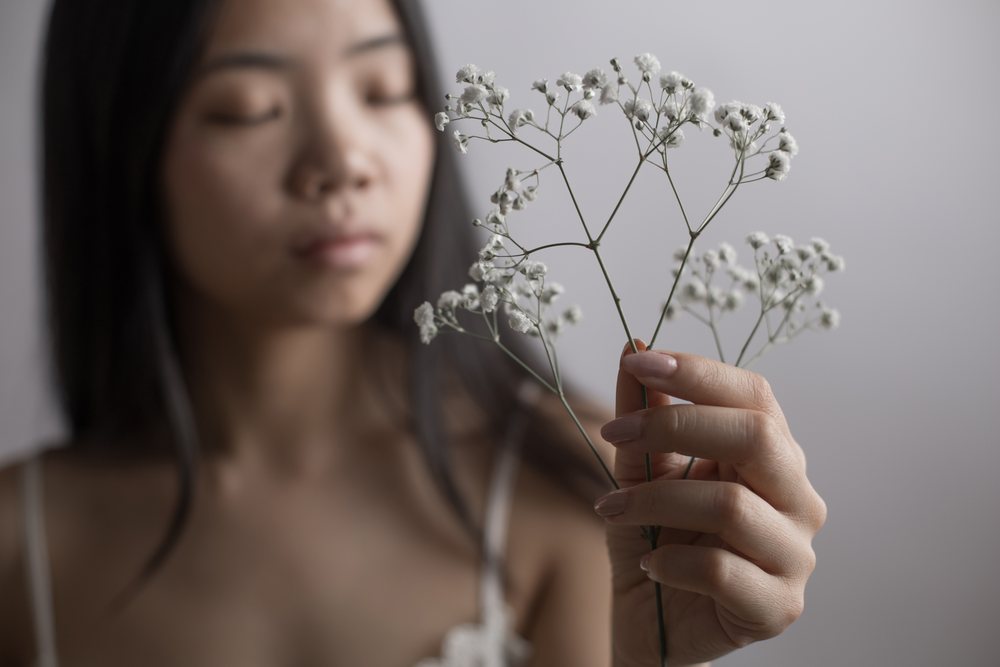 Young woman with closed eyes holding delicate white baby's breath flowers in soft natural light against a gray background, Art, portrait, emotional.