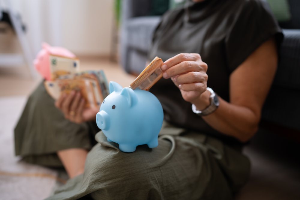 Person sitting indoors on floor inserting euro banknote into blue piggy bank on lap with multiple currency notes and a pink piggy bank nearby, Spain.