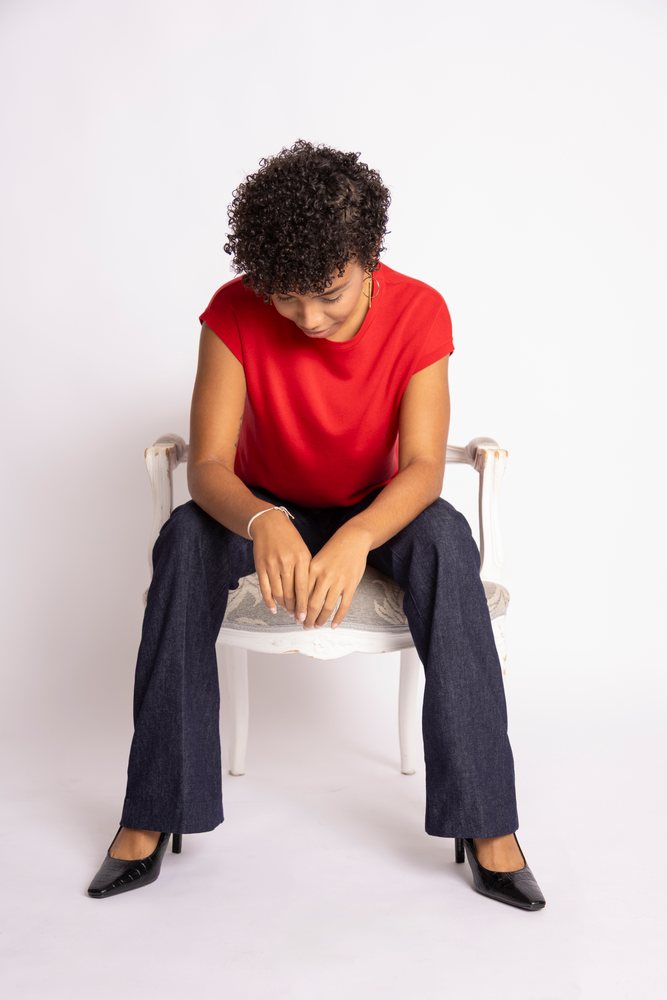 Woman sitting on a modern chair looking down, wearing a red shirt, blue pants, and black high heels in a studio, fashion, clothes, young culture.