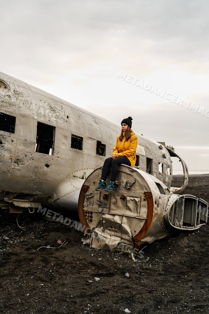 A white woman sits on airplane wreckage on a black sand beach