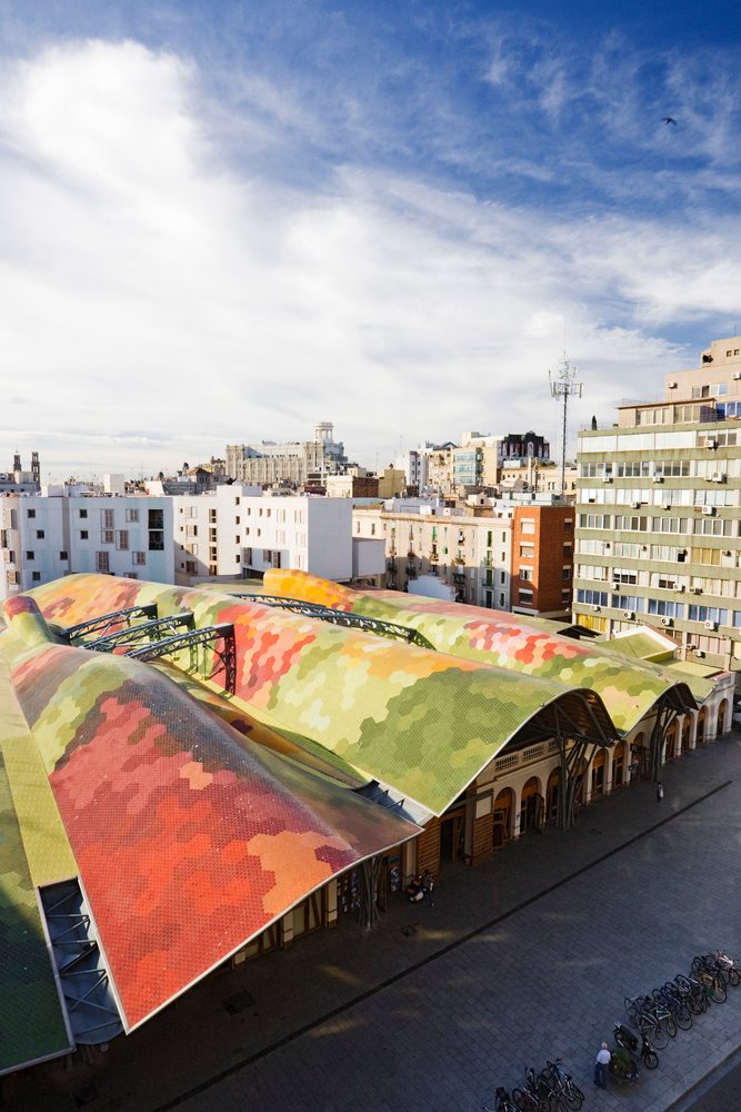 Santa Catarina Market features a colorful hexagonal tiled roof with people walking near bicycles on a pedestrian street under a partly cloudy sky, Catalunya, Spain.