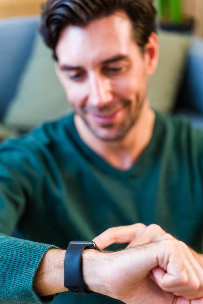 Smiling man in green sweater touching black smartwatch on his wrist indoors, Spain.