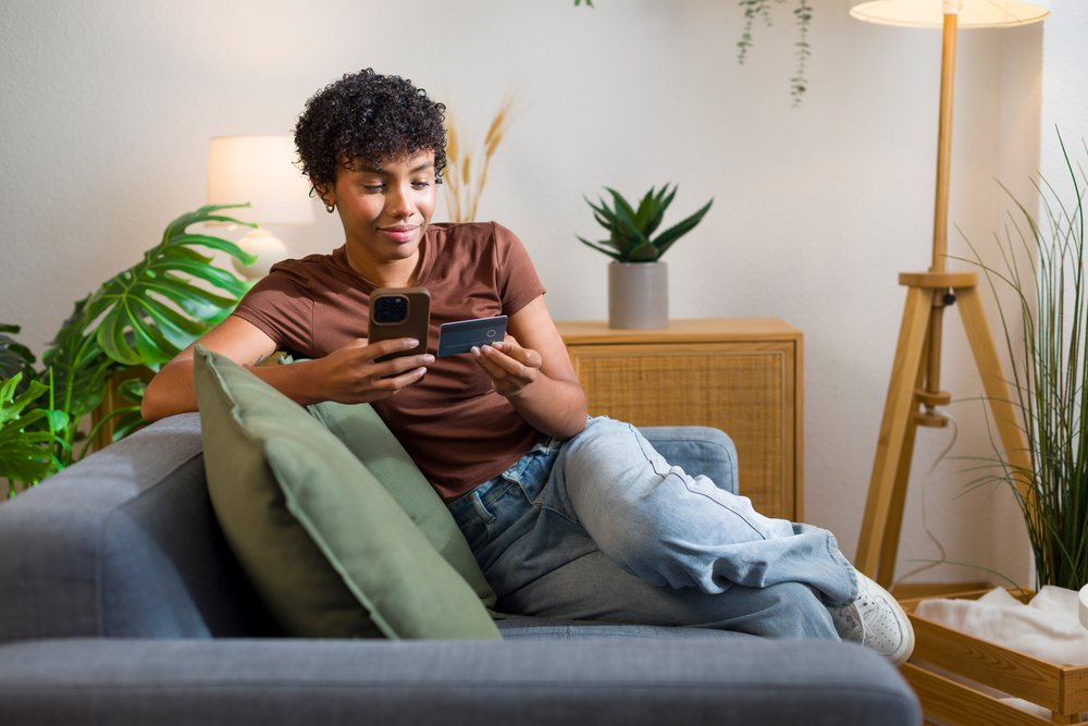 Young woman seated on a couch holding a smartphone and credit card in a modern home living room.