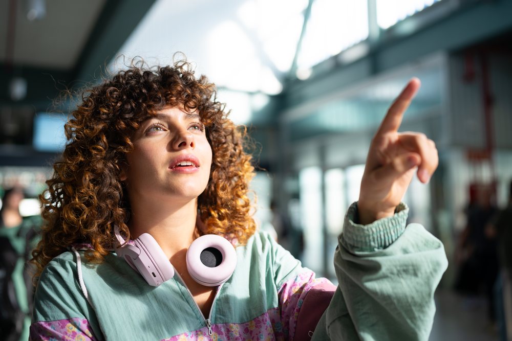 Woman with curly hair wearing over-ear headphones points inside a bright modern building with glass walls, Spain.