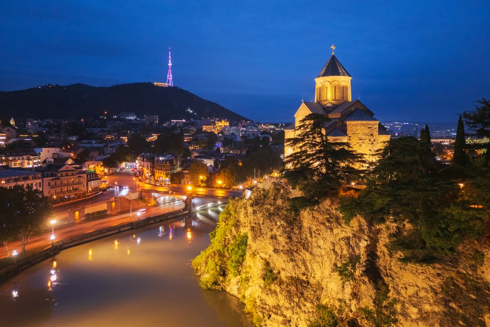 Metekhi Church illuminated on a rocky cliff above the Mtkwari River at night with city lights and hills in the background, Cityscapes, Tbilisi, Georgia.