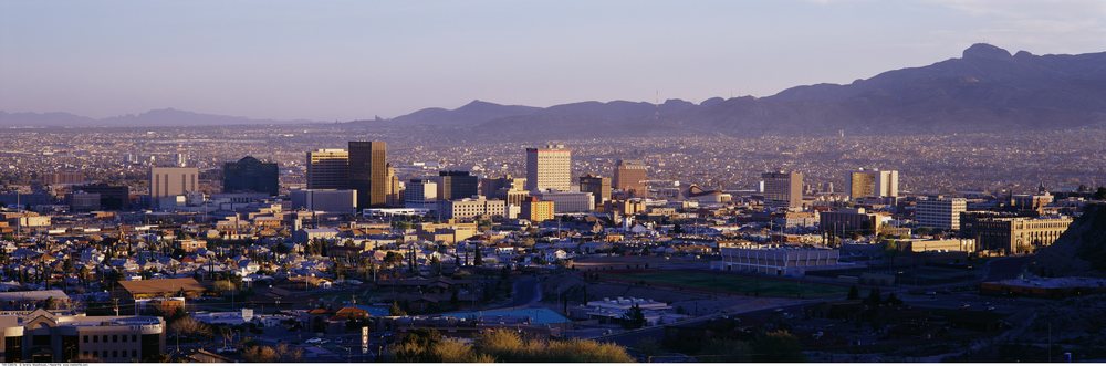 Panoramic cityscape of El Paso, Texas, showing downtown buildings and mountains at sunset with clear sky and sunlight casting shadows.