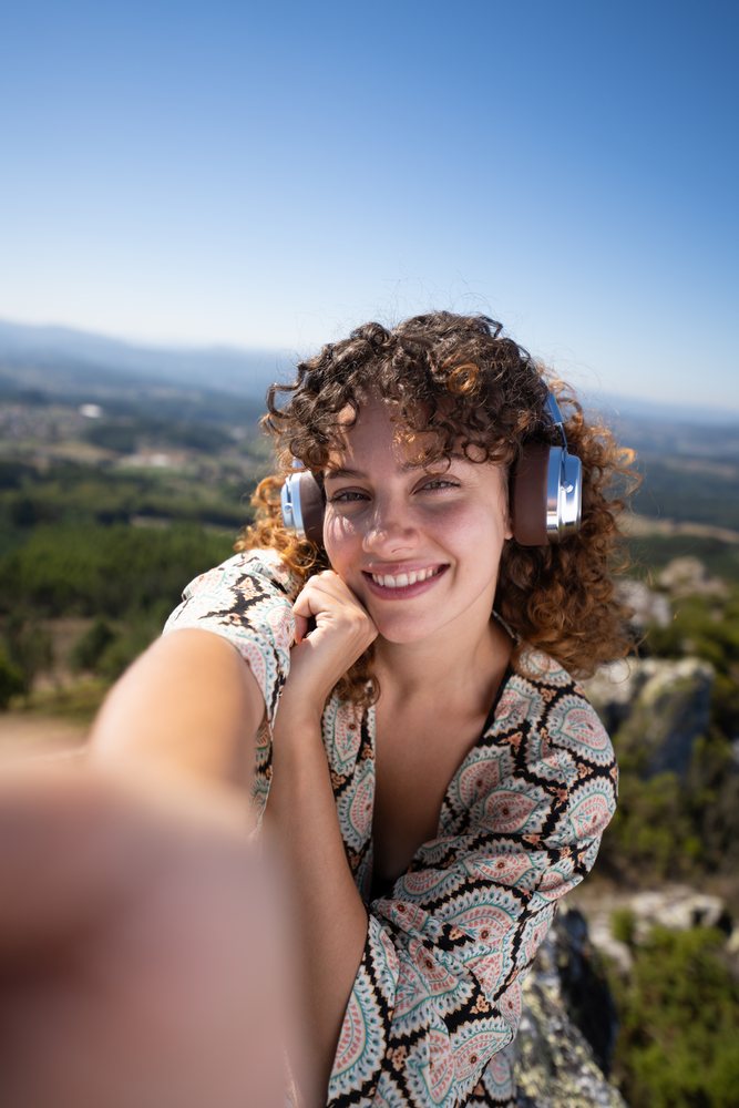 Woman with curly hair wearing headphones takes a selfie outdoors with the scenic Pico Sacro mountain landscape, Spain behind her, Galicia.