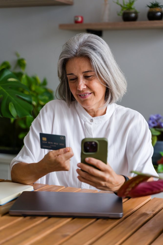 Mature woman with gray hair holding a card and using a smartphone at a wooden table with a laptop and plants indoors, Spain.