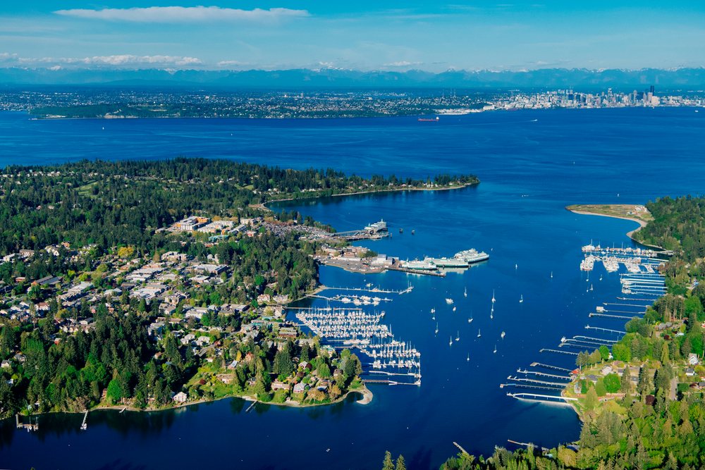 Aerial view of Bainbridge Island and Eagle Harbor showing boats docked at the marina, ferries, and city skyline across Puget Sound under clear skies, Salish Sea, USA.
