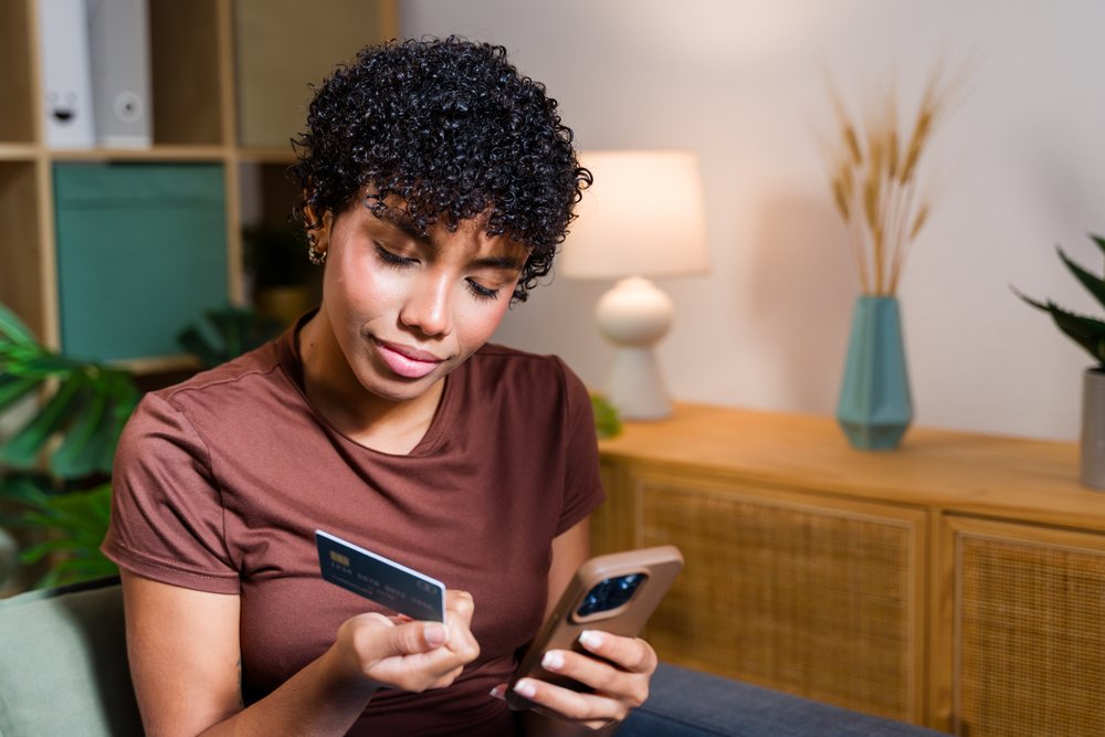 Young woman a credit card and a mobile phone while seated in a living room with plants and a lamp on a wooden cabinet, Spain.