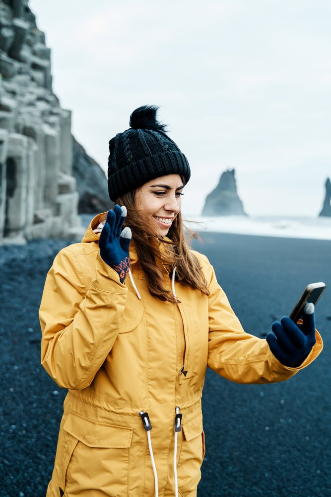 A white woman uses her phone for a video call on a black sand beach