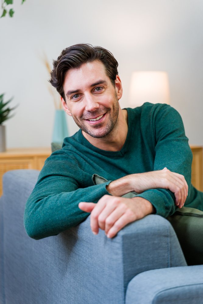 Man in a green sweater smiles while sitting on a sofa in a living room with a lamp and plant in the background, Spain.