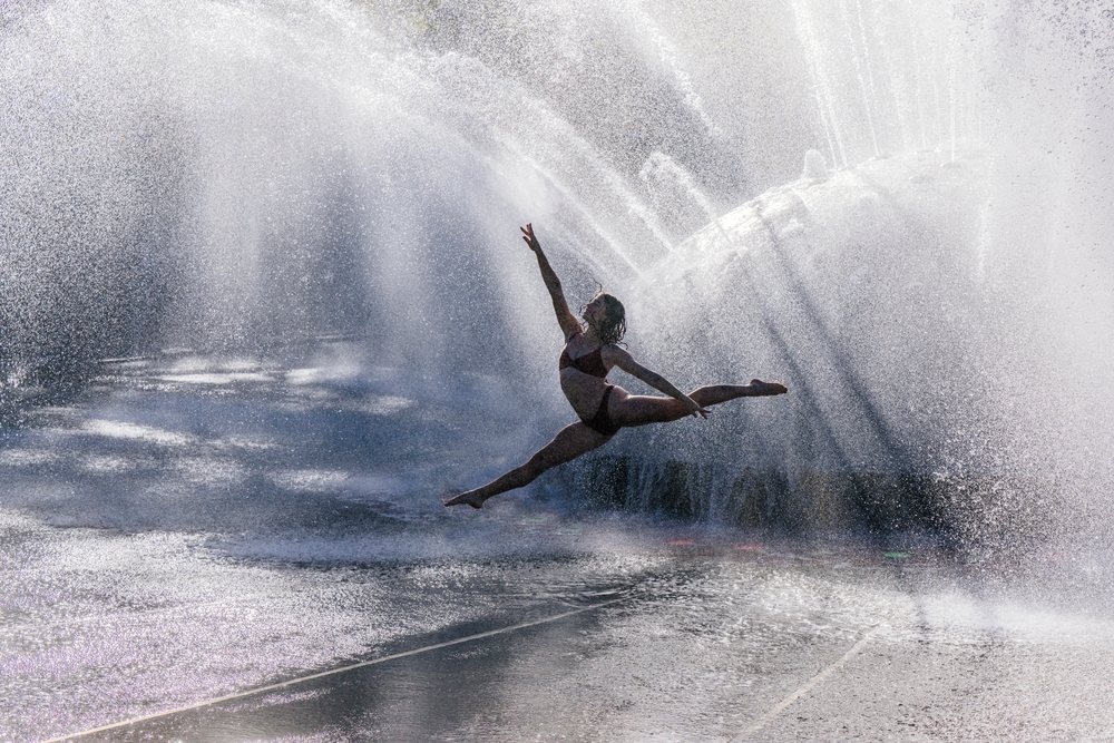 Woman in black bikini performing a graceful dance leap through a water fountain spray outdoors, partner, acrobatics, yoga, USA.