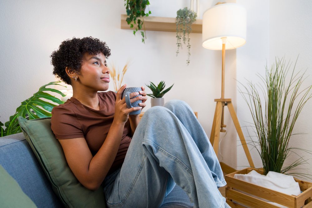 Young woman with curly hair sits on a gray sofa holding a blue mug amid indoor plants and a floor lamp in a cozy home.