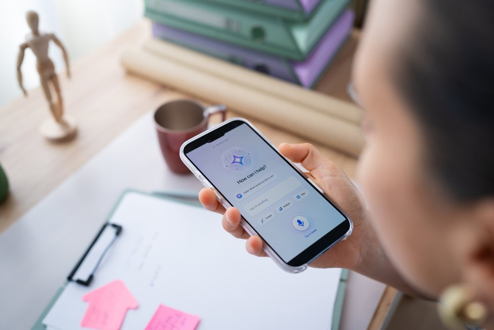 Person holding smartphone with voice assistant app at a desk with clipboard, sticky notes, coffee mug, and wooden mannequin, telecommuting, Spain.