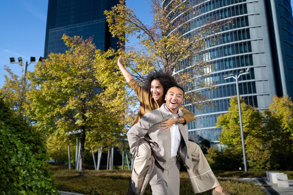 Man giving woman a piggyback ride in a sunny urban park with autumn trees and modern glass buildings, Madrid, Spain.
