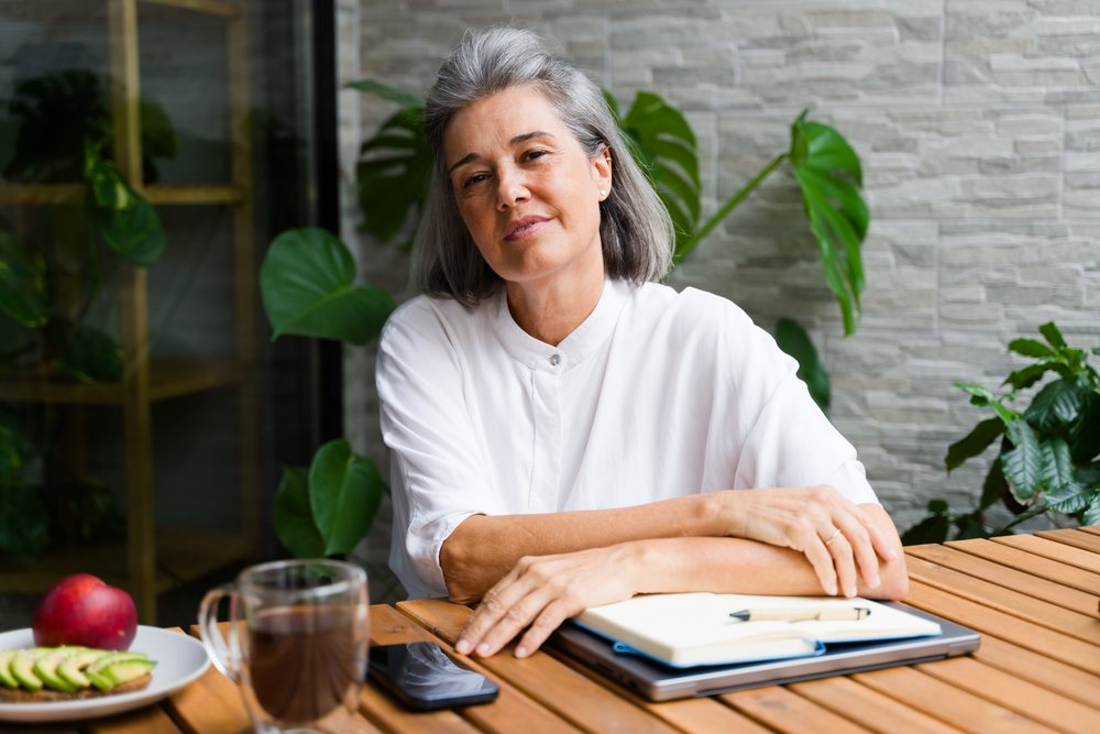 Middle-aged woman sitting at a wooden table with a notebook, pen, laptop, smartphone, coffee cup, and avocado toast, surrounded by indoor plants and a tile wall, Spain.
