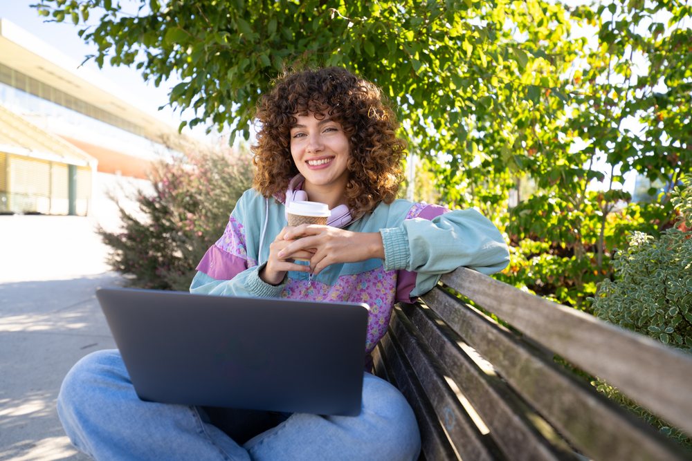 Woman with curly hair sits on a bench outdoors smiling while holding a coffee cup and using a laptop on a sunny day, Spain.