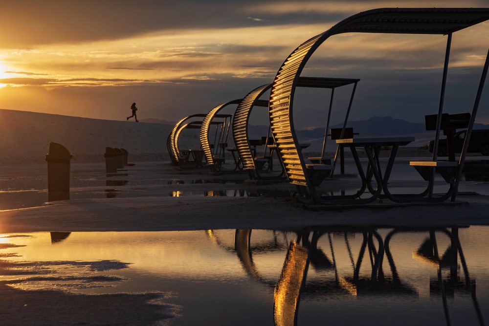 Picnic shelters with curved roofs reflect in water puddles near a sand dune where a runner is visible at Sunset in white Sands National Monument, Landscape, Wildlife, Alamogordo, New Mexico, USA.