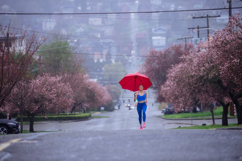 Woman running on a rainy urban street holding a red umbrella amidst blooming pink blossom trees in spring, USA.