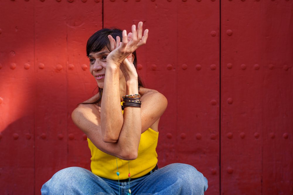 Woman in a yellow top and blue jeans sits relaxed outdoors against a red door, under sunlight, retro, vintage, Málaga, Spain.
